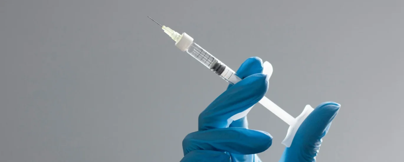 Gloved hand holding a syringe with a visible needle, against a neutral gray background, symbolizing medical procedures or vaccinations.